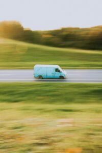 A blue van speeds through the countryside along a sunlit road, capturing motion in the summer landscape.
